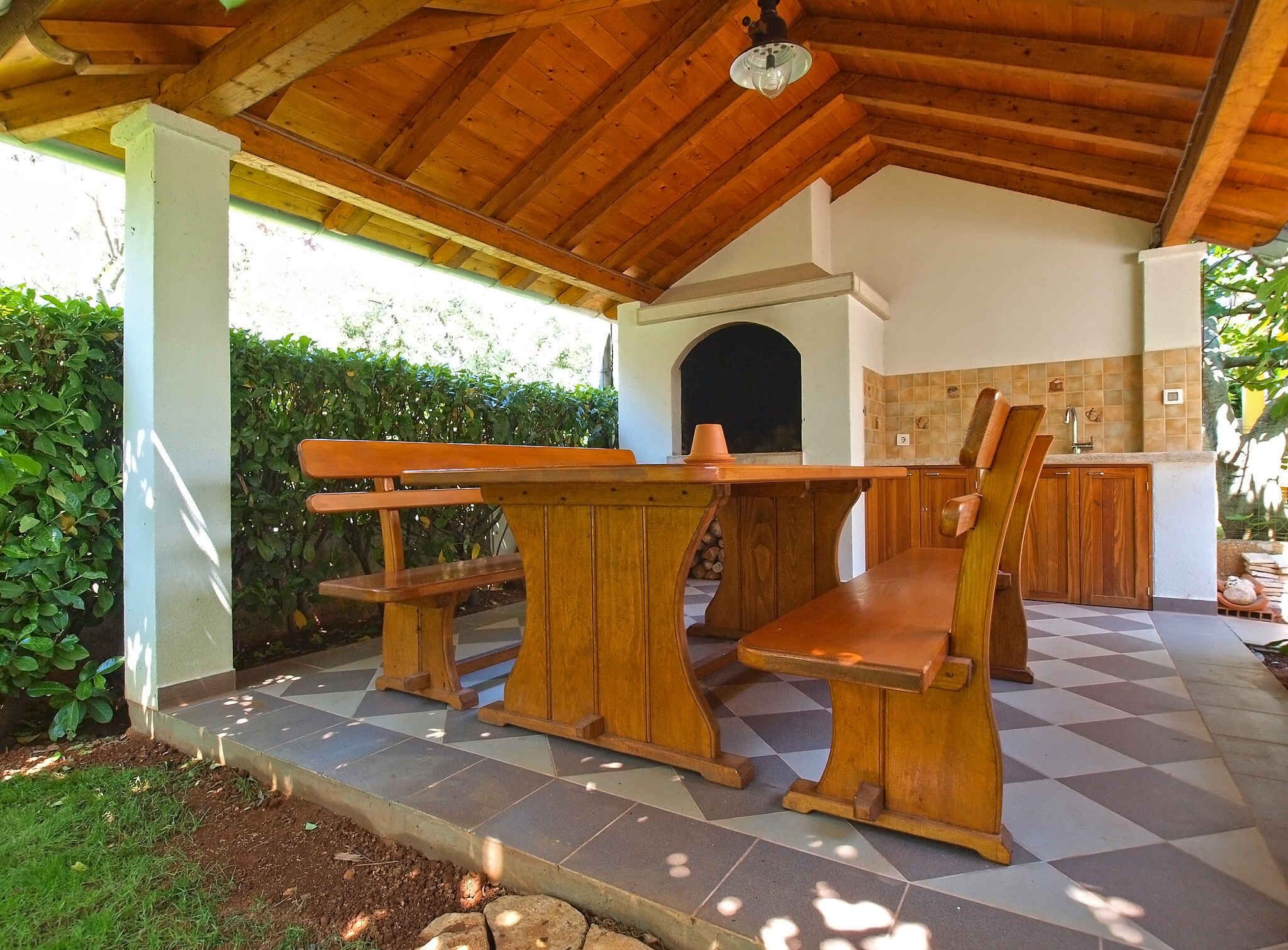 An outdoor patio with a wooden table and benches, a tiled floor, and a kitchen area with cabinets and a sink.