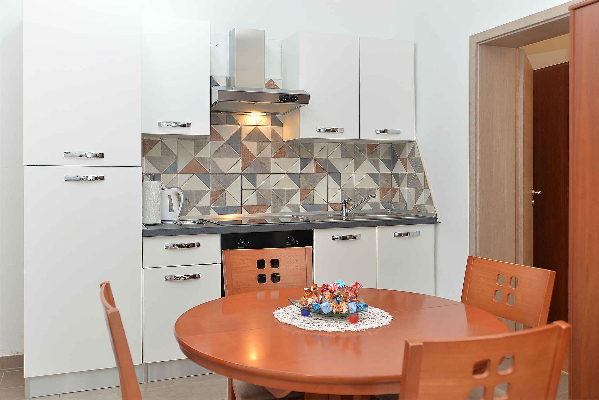 A compact kitchen with white cabinets, a tiled backsplash, and a round wooden dining table with chairs.