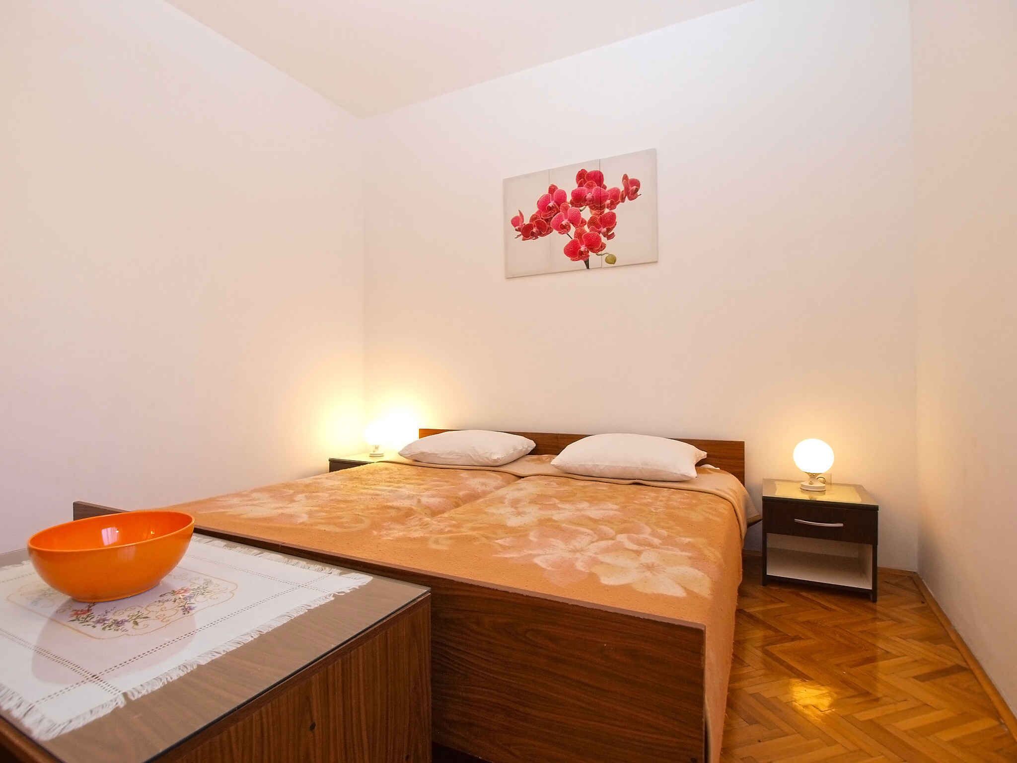 Une chambre avec un lit en bois, deux tables de chevet, une petite table, un bol posé sur une table et un tableau floral accroché au mur.