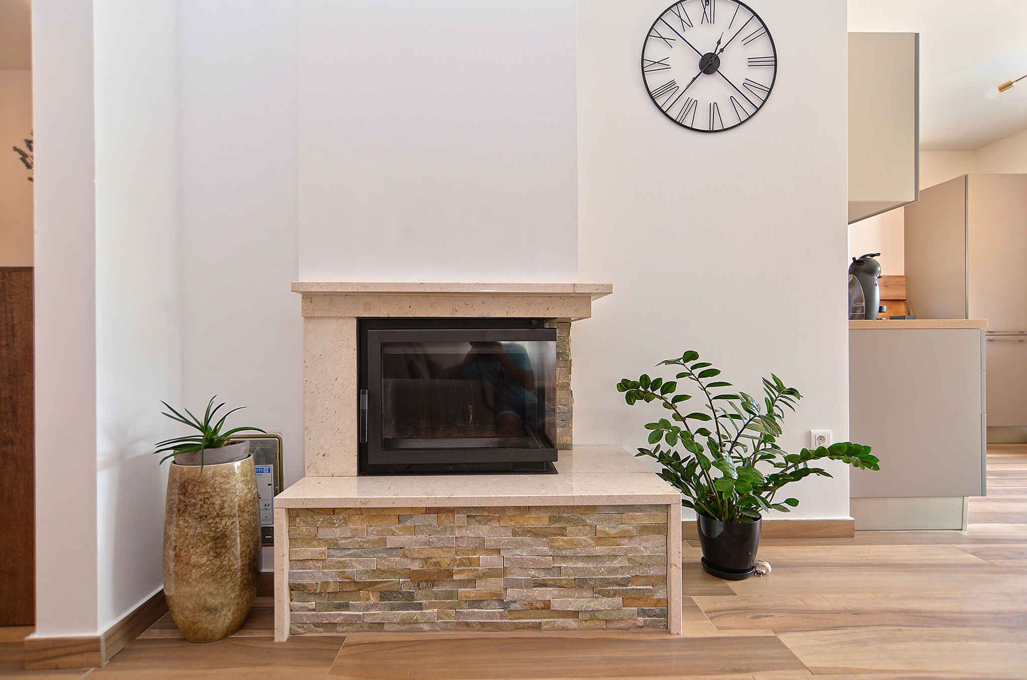 A modern living room with a stone fireplace, a large wall clock, and potted plants.