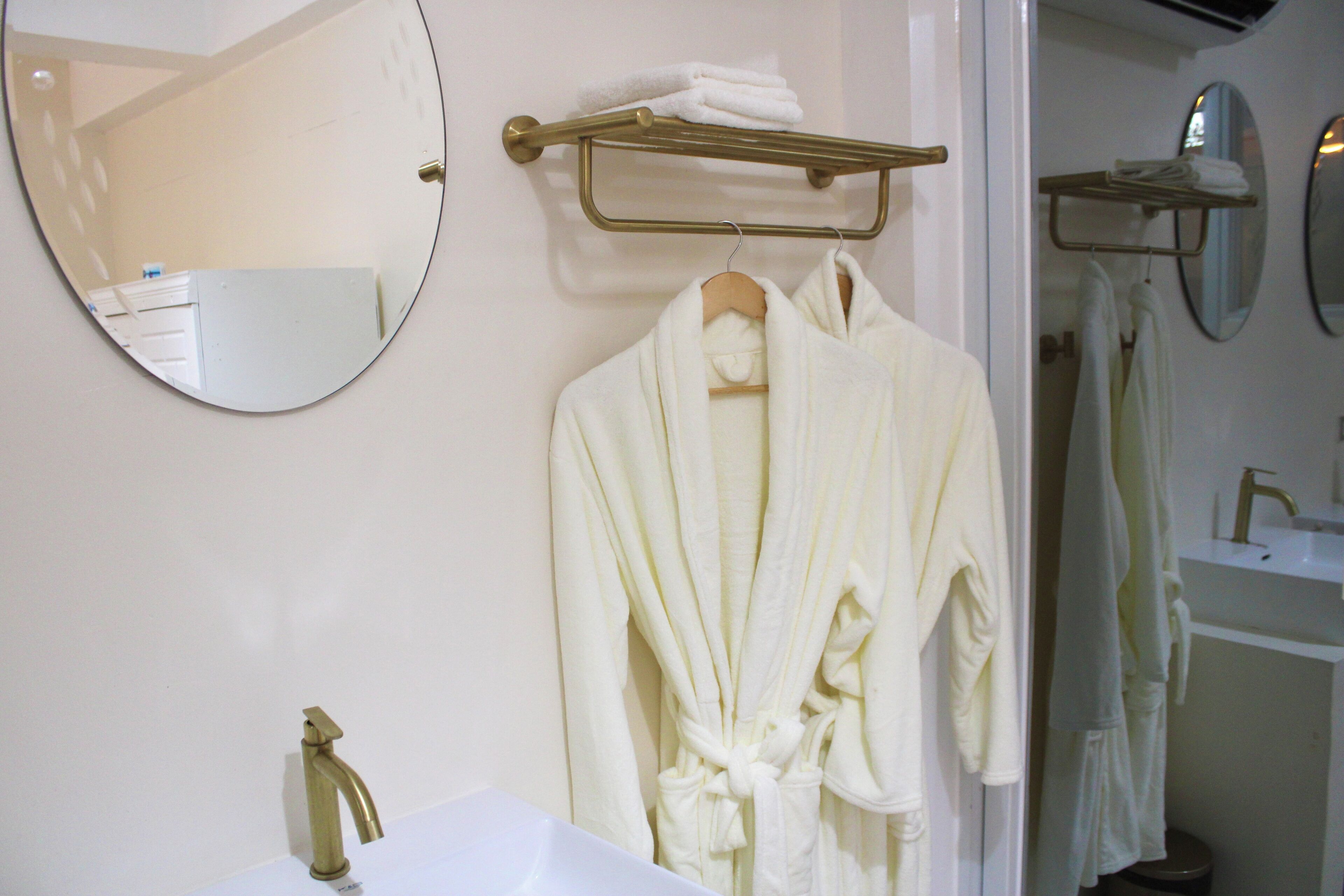 Un baño con un espejo redondo, un toallero dorado, un lavabo blanco y una bata blanca colgada de un perchero de madera.