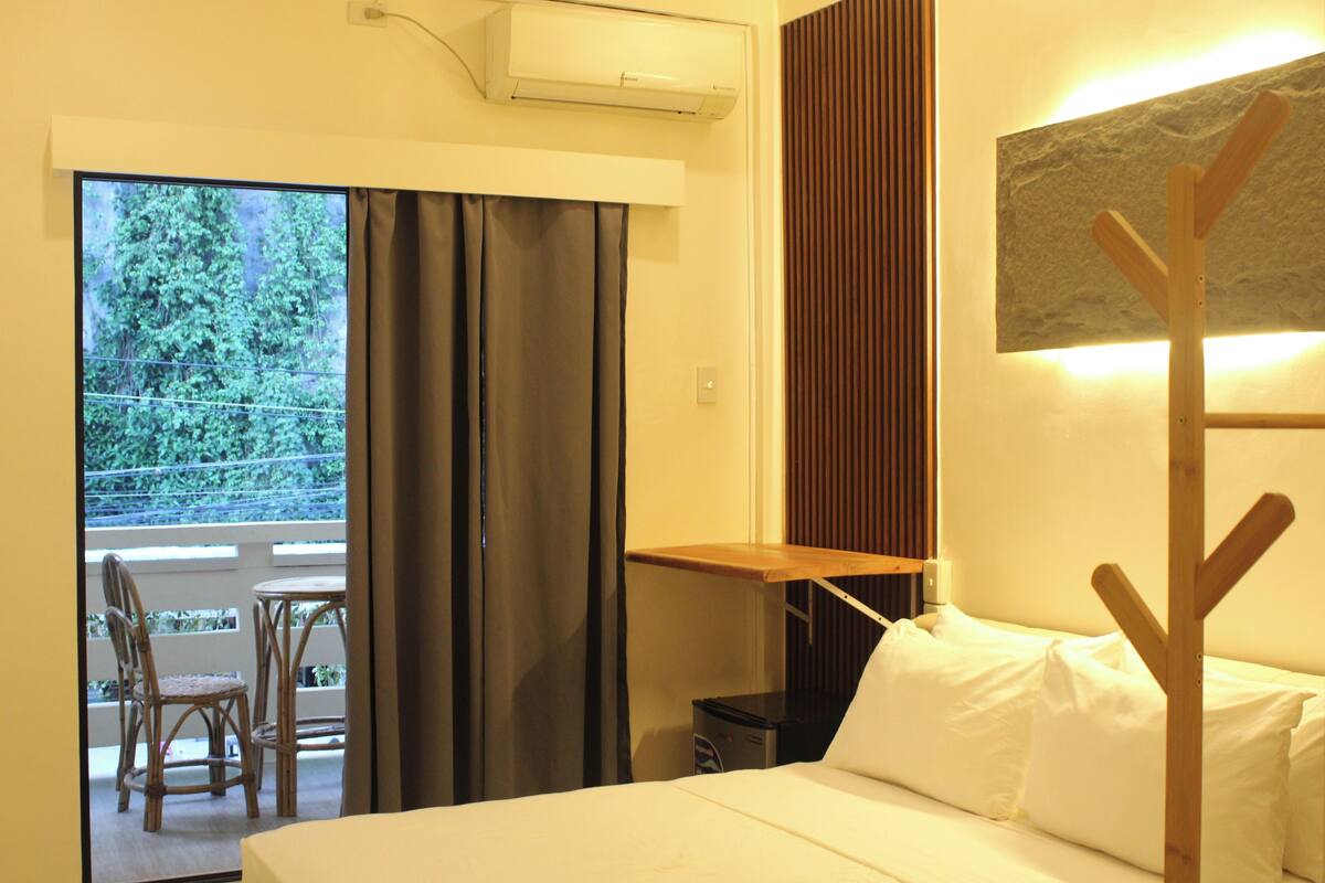 A hotel room with a bed, a small table, a chair, and a view of greenery through the window.
