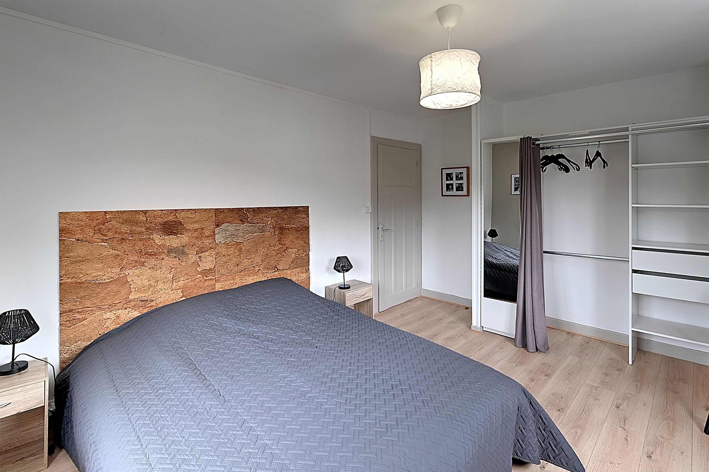 Une chambre avec un grand lit, une tête de lit en bois, une table de chevet avec une lampe, une armoire et une porte donnant sur une autre pièce.