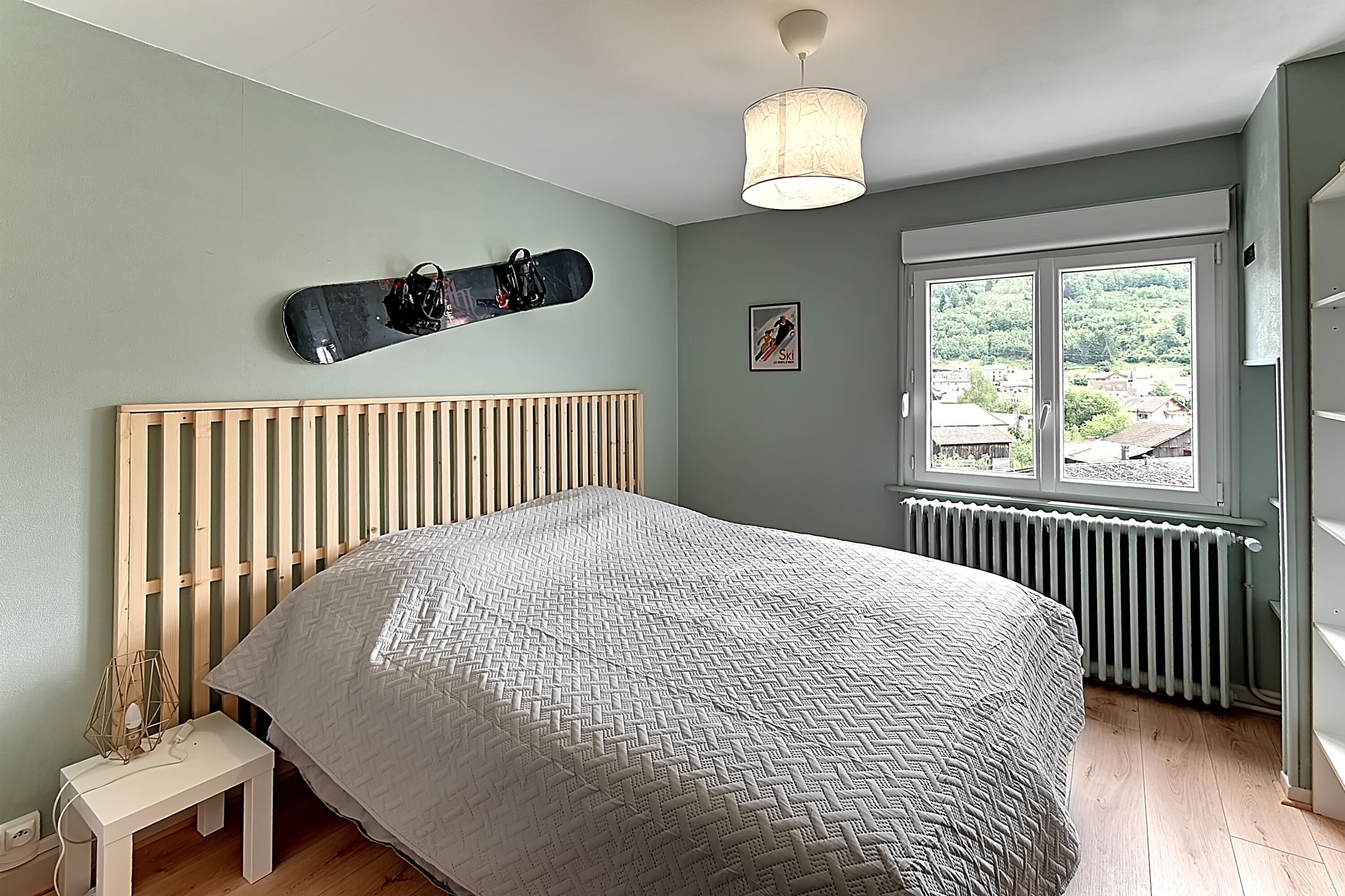 A bedroom with a bed, a wooden headboard, a window with a view, a wall-mounted snowboard, and a small table with a lamp.