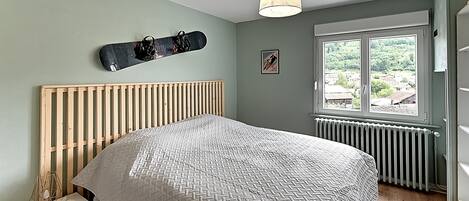 A bedroom with a bed, a wooden headboard, a window with a view, a wall-mounted snowboard, and a small table with a lamp.