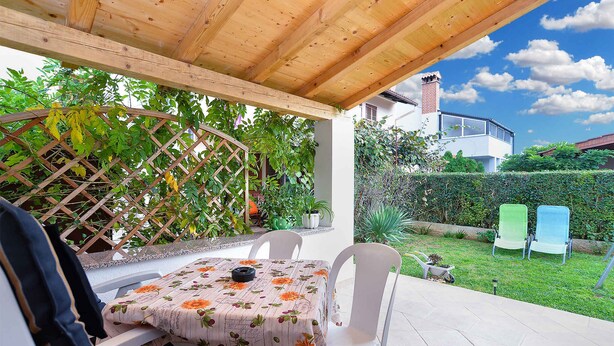 Un patio cubierto con una mesa y sillas, rodeado de follaje y una casa al fondo.