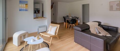 A modern living room with a sofa, a small table, and a chair. There is a dining area with a table and chairs in the background.