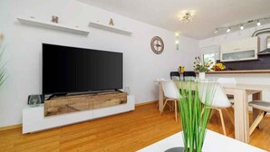 Una sala moderna con televisor de pantalla plana, piso de madera, un comedor con mesa y sillas blancas, y una cocina con microondas y armarios.