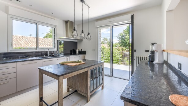 A modern kitchen with a central island, large windows, and a sliding glass door leading to a balcony. - Villa Violette - Piscine privée (Gujan-Mestras)
