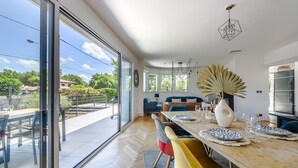 A dining area with a large table, chairs, and a view through glass doors to an outdoor patio. - Villa Violette - Piscine privée (Gujan-Mestras)