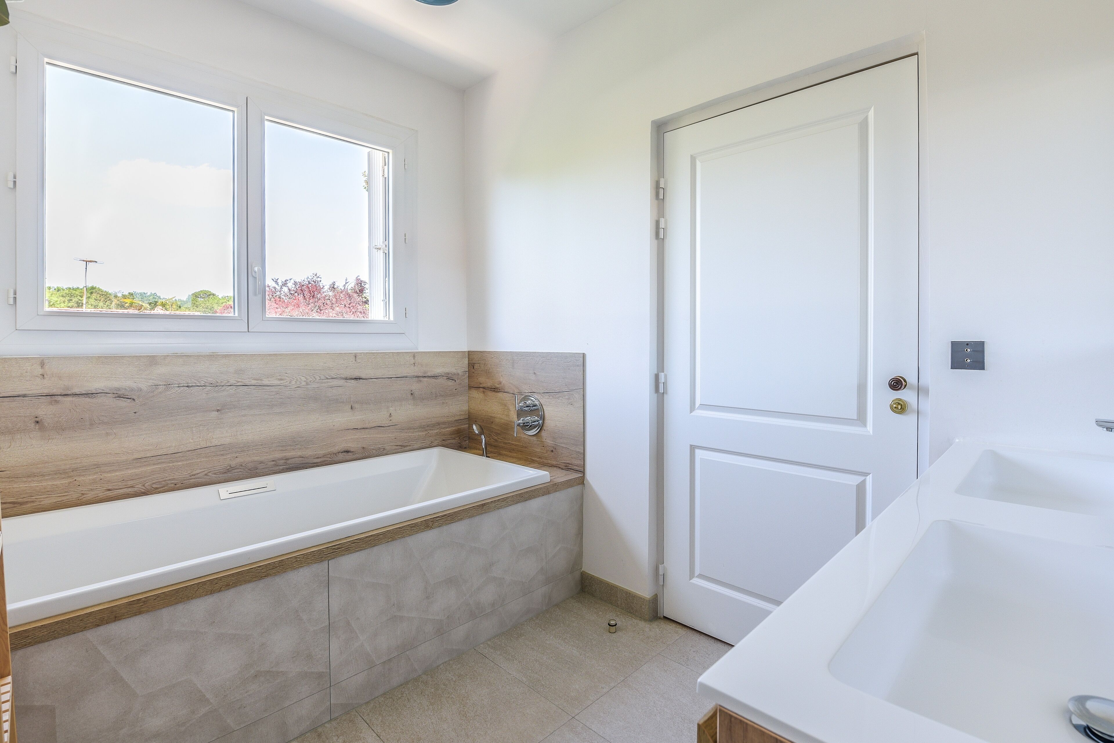 A modern bathroom with a freestanding bathtub, a window with a view of greenery, and a white door.