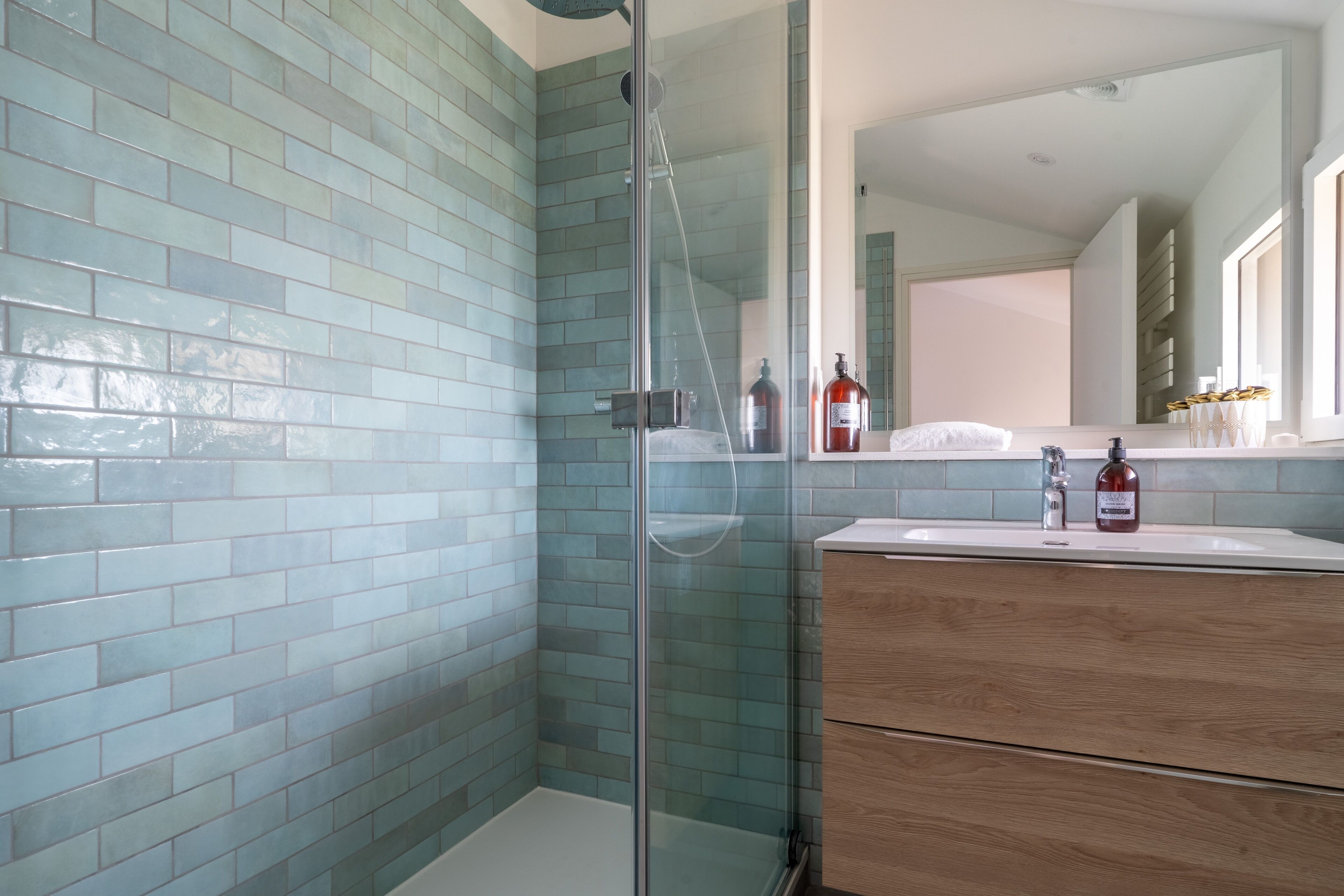 Ein modernes Badezimmer mit blauen Fliesen, einer Glasdusche, einem Holzkosmetikschrank und einem großen Spiegel.