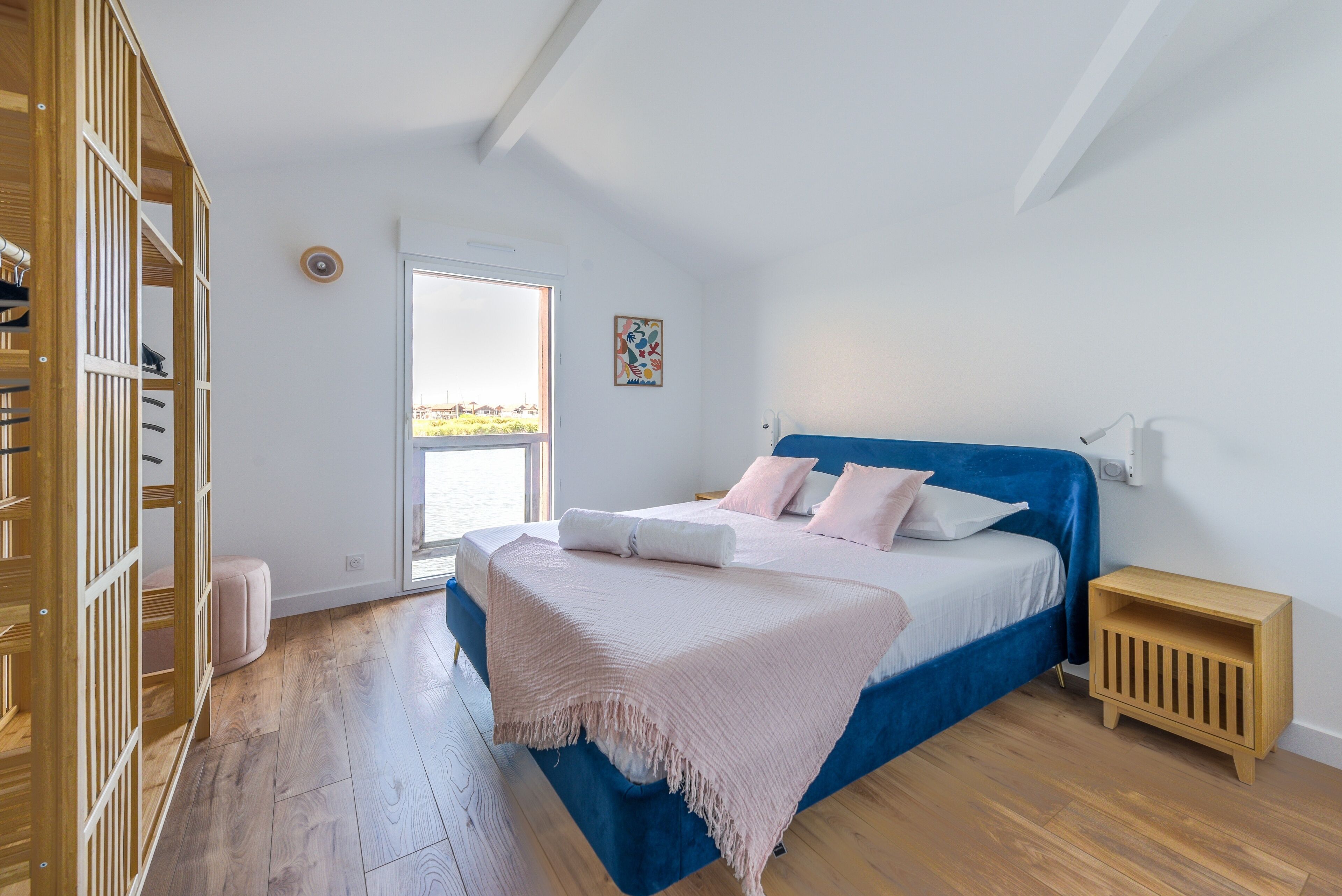 A bedroom with a large bed, a wooden wardrobe, a small bedside table, and a window with a view.