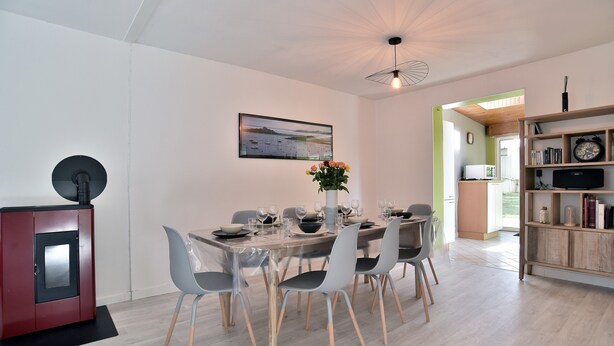 A modern dining area with a wooden table set for six, surrounded by grey chairs. A red cabinet with a black circular object is to the left. A framed picture hangs on the wall above the table.