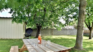 A wooden picnic table with six glasses and a bottle of drink, set in a grassy backyard with trees and a barbecue grill in the background.
