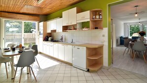 A modern kitchen with white cabinets, a wooden countertop, and a dishwasher. There is a dining area with a table and chairs, and a view of the outdoors through large windows.