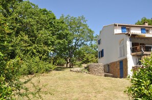 Un bâtiment blanc aux volets bleus, entouré d’arbres et d’un mur de pierre.