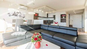 A modern living room with a black leather sectional sofa, a white coffee table with a red vase, and a kitchen area in the background.