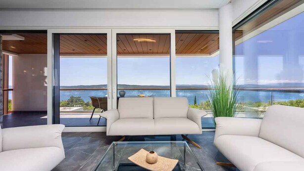 En moderne stue med et glasbord, en beige sofa og udsigt til vand gennem store vinduer.