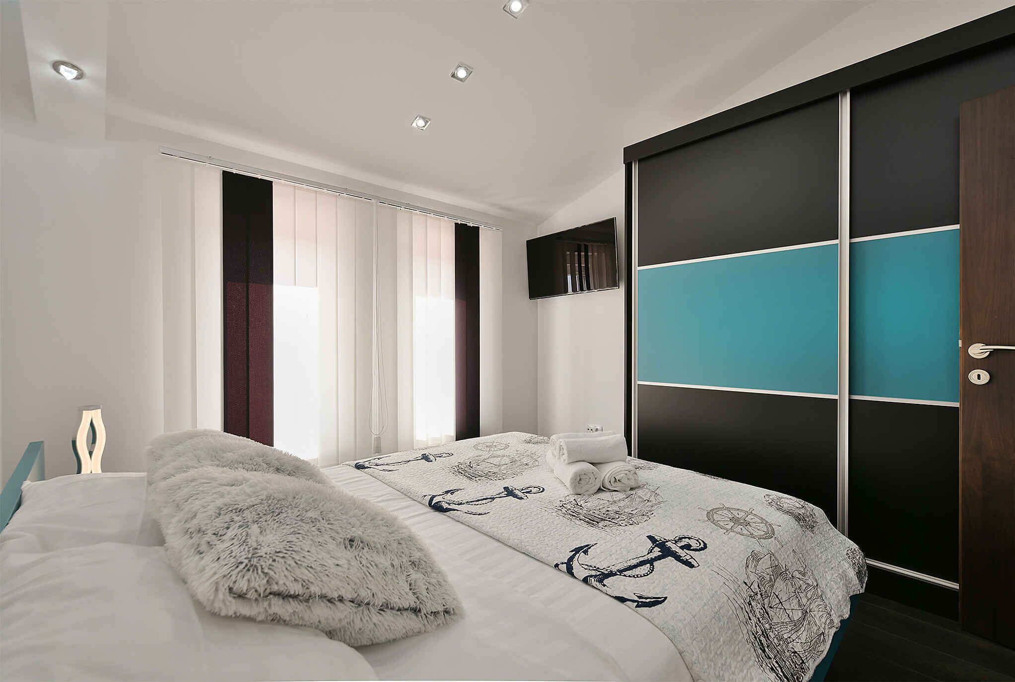 Quarto moderno com cama, uma janela grande com cortinas, TV fixada na parede e guarda-roupa com painel decorativo azul-esverdeado.
