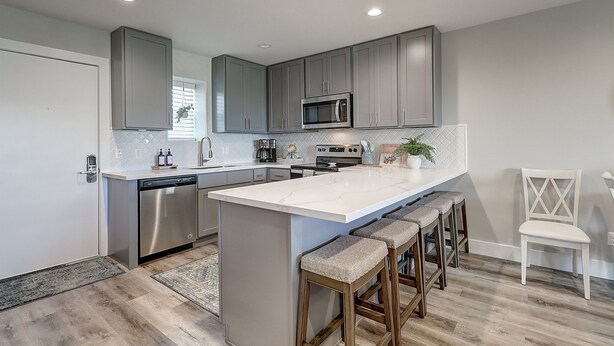 A modern kitchen with grey cabinets, stainless steel appliances, a large island with bar stools, and a white chair. - Beachfront Luxury with Gulf Views: 3BR/2BA Condo w/ Private Beach Access & Pool (Port Aransas)
