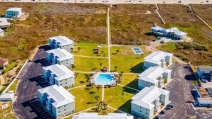 An aerial view of a coastal hotel with multiple buildings, a central swimming pool, and a beach in the background. - Beachfront Luxury with Gulf Views: 3BR/2BA Condo w/ Private Beach Access & Pool (Port Aransas)