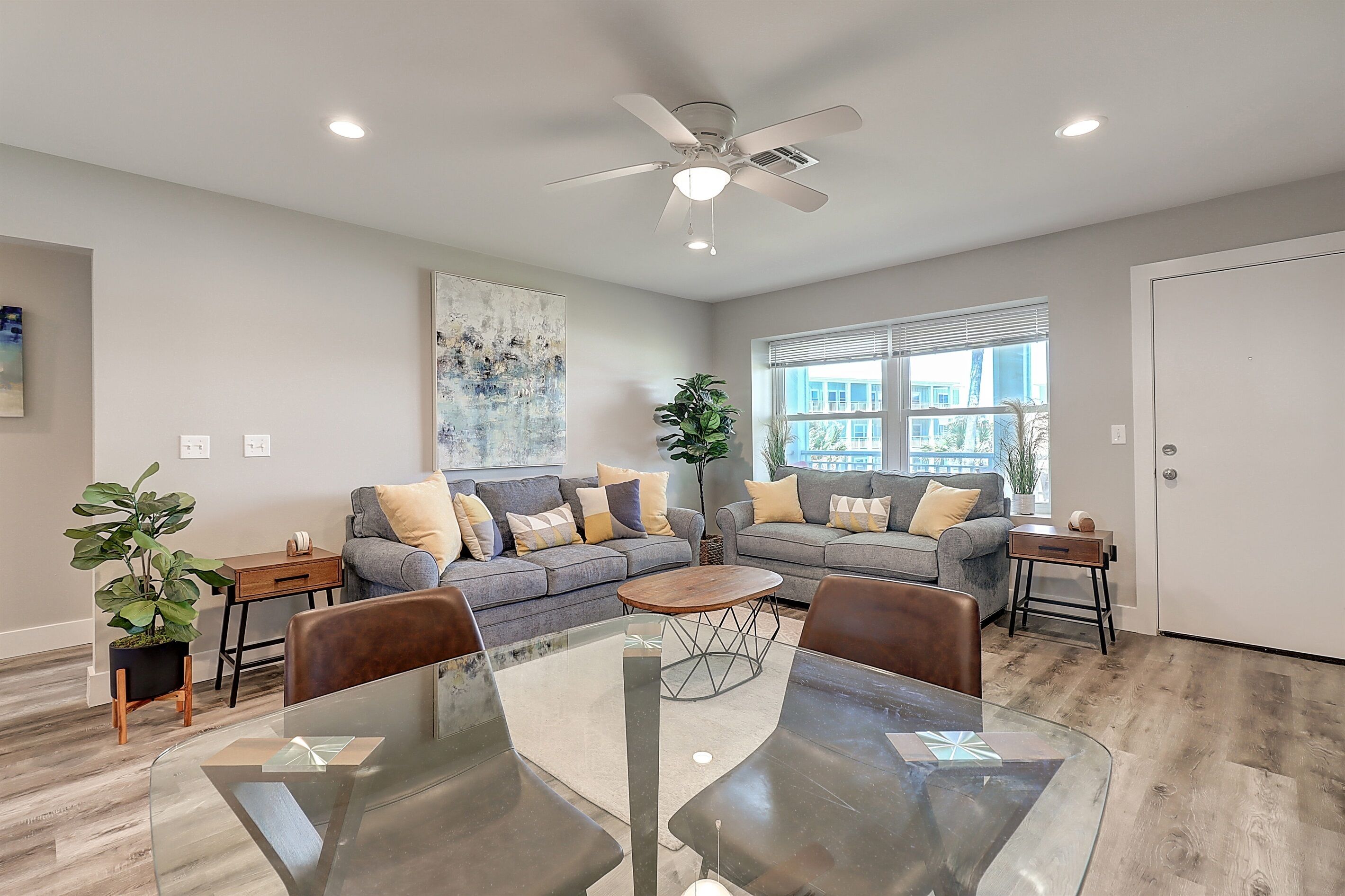 A modern living room with a glass dining table, two grey sofas, a ceiling fan, and a large window.