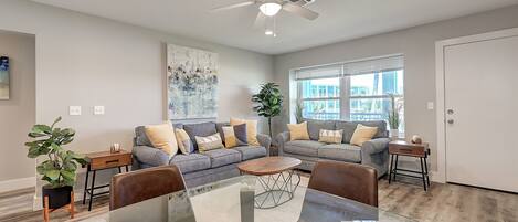 A modern living room with a glass dining table, two grey sofas, a ceiling fan, and a large window.