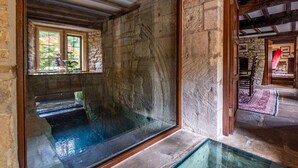 A stone-walled room with a glass-bottomed pool, wooden beams, and a view of a garden through a window. - Midsummer Mill - Five Bedroom Apartment, Sleeps 8 (Bourton-on-the-Water)