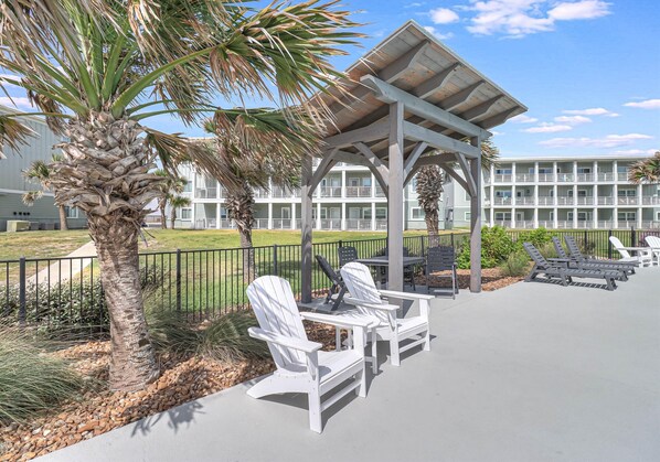 A covered outdoor seating area with Adirondack chairs, a palm tree, and a building with balconies in the background. - Remodeled Beachy 2BR/2BA-Pool View & Walk to Beach (Port Aransas)