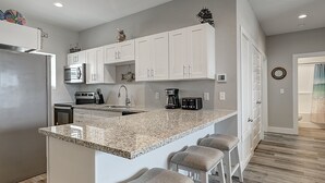 Eine Küche mit weißen Schränken, einer Arbeitsplatte aus Granit, einem Edelstahlkühlschrank, einer Mikrowelle, einer Kaffeemaschine und einem Spülbecken.