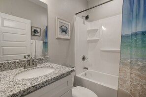 A bathroom with a granite countertop sink, a white toilet, a bathtub, and a shower curtain depicting a beach scene. - Newly Remodeled Beachy 2BR/2BA Condo with Pool View and Close Walk to the Beach (Port Aransas)