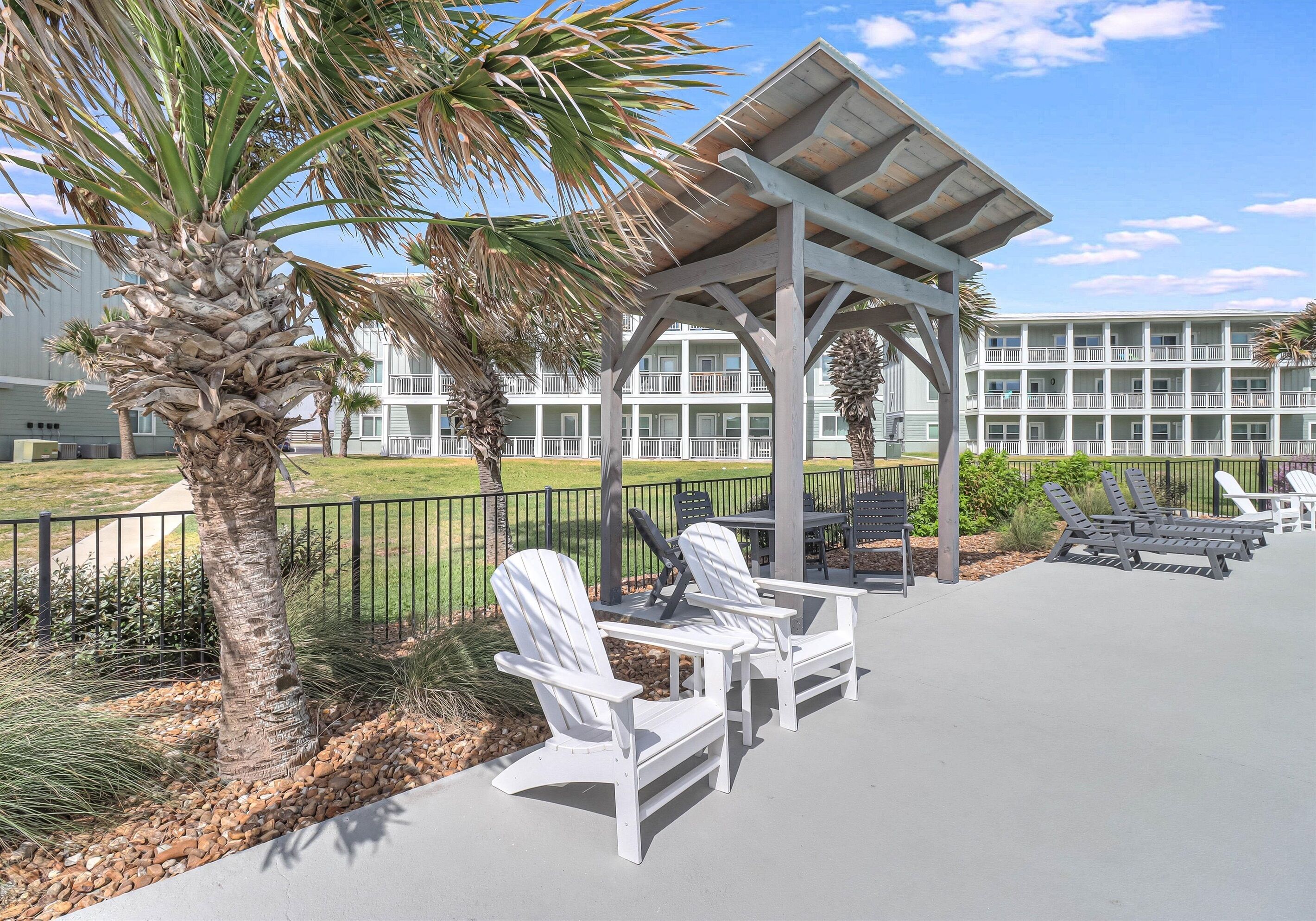 A covered outdoor seating area with Adirondack chairs, a palm tree, and a building with balconies in the background.