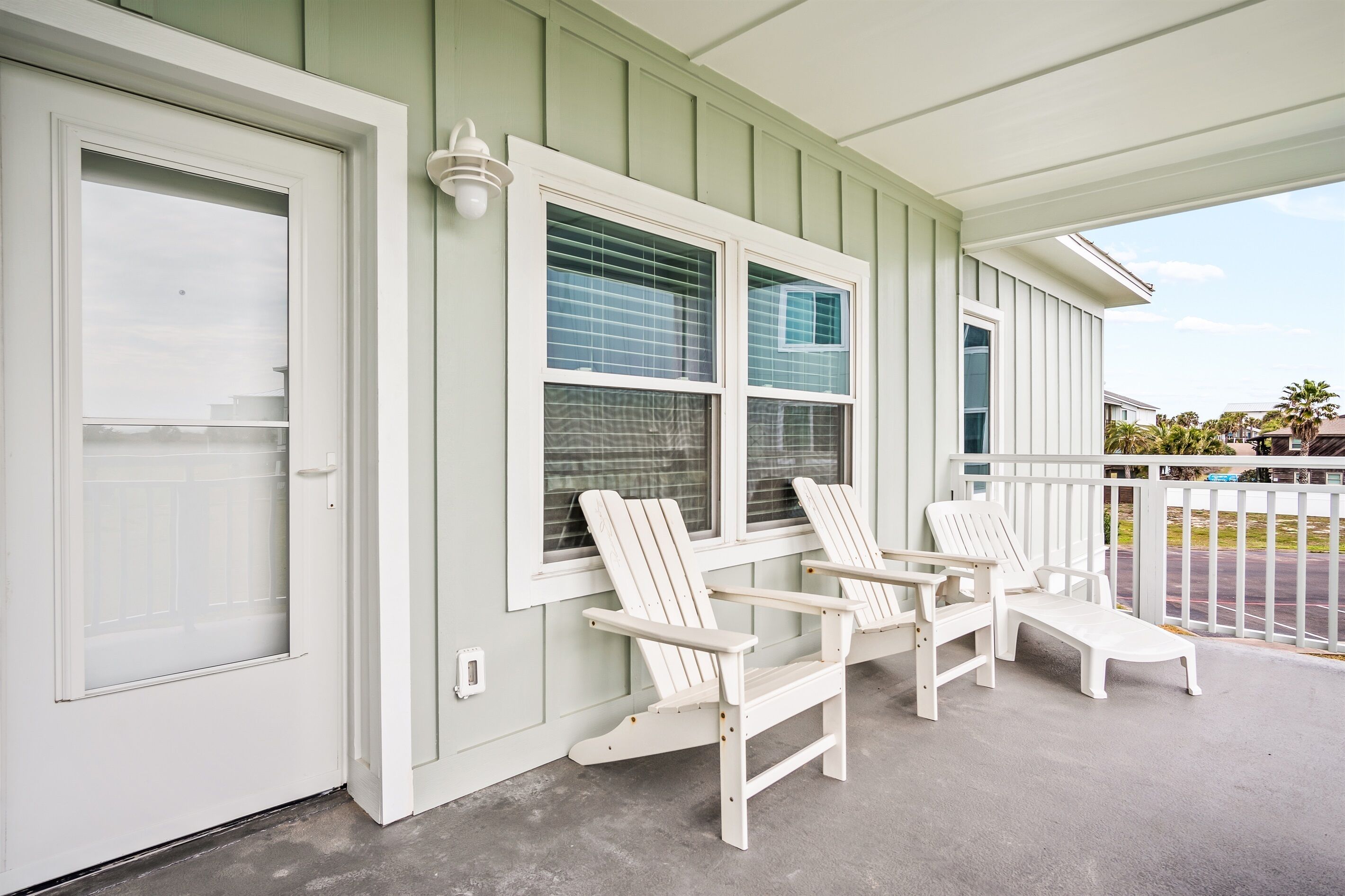A porch with two Adirondack chairs and a lounge chair, a white door, and a window with blinds.