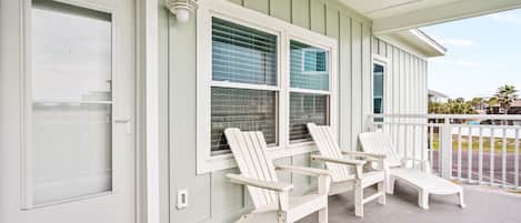 A porch with two Adirondack chairs and a lounge chair, a white door, and a window with blinds.