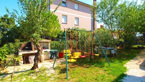A backyard with a swing set, a barbecue grill, and a table with chairs. - Holiday apartment with garden for shared use and children's playground (Ližnjan)