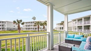 A covered balcony with wicker furniture and a view of adjacent buildings and palm trees. - Remodeled 3BR Condo: Private Beach Access, Pool View, & Walking Distance to Town (Port Aransas)