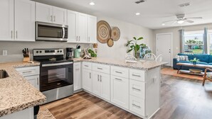 A modern kitchen with white cabinets, a stainless steel oven, and a microwave. There is a dining area with a blue sofa and a ceiling fan. - Remodeled 3BR Condo: Private Beach Access, Pool View, & Walking Distance to Town (Port Aransas)
