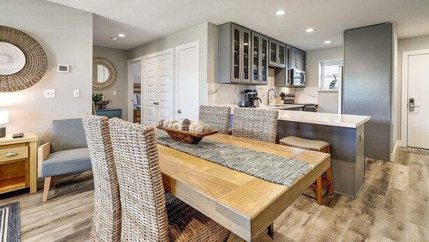 A modern kitchen with a central island, grey cabinets, and a dining area with wicker chairs and a wooden table. - Newly Remodeled 2BR Condo with Private Path to Beach, Pool View, & Close to Town (Port Aransas)