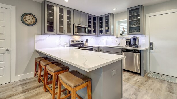 A modern kitchen with grey cabinets, a large island with stools, stainless steel appliances, and a clock on the wall. - 3BR Condo w/ Pool View, Private Beach Access & Short Walk to Town and the Beach (Port Aransas)