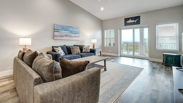 A spacious living room with a large sofa, a coffee table, and a ceiling fan. There are two windows with blinds, a door leading to a balcony, and a painting on the wall. - Luxury Gulf-View Penthouse – Top Floor 3BR Condo (Port Aransas)