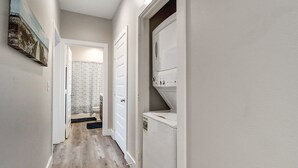A hallway with a wooden floor, a door leading to a bathroom, and a laundry machine on the right. - Luxury Gulf-View Penthouse – Top Floor 3BR Condo (Port Aransas)