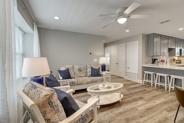 Relax in this stylish living room with a sofa set, round coffee table, and ceiling fan, adjacent to a modern kitchen with white cabinetry and bar counter.