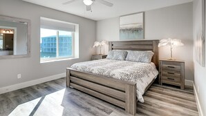 Une chambre à coucher avec un lit, une tête de lit en bois, des tables de chevet, un ventilateur de plafond, une fenêtre et un tableau au mur.