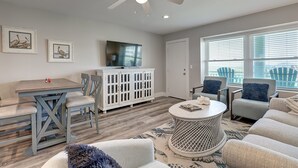 A living room with a dining area, a ceiling fan, a television, and a large window with blinds. - Experience Paradise in this Recently Remodeled 3BR/2BA (Port Aransas)