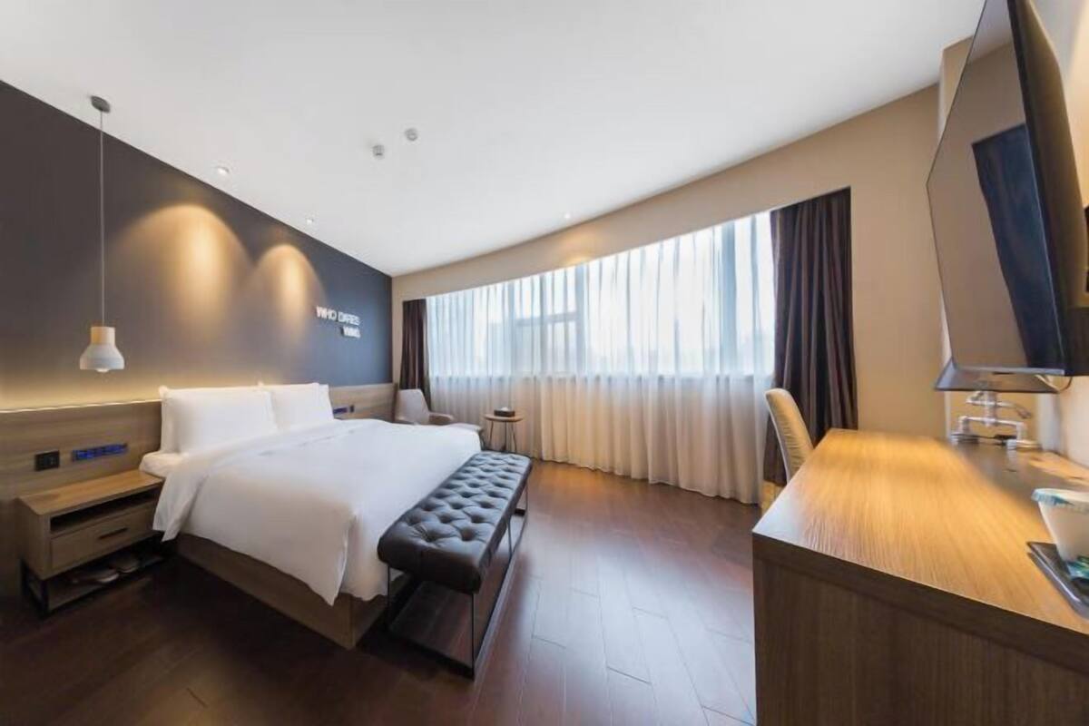 Une chambre d’hôtel moderne avec un grand lit, un bureau, une banquette et une grande fenêtre avec des rideaux.