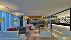 Um lobby moderno de hotel com balcão de recepção, área de estar e vista para o horizonte urbano através de amplas janelas.