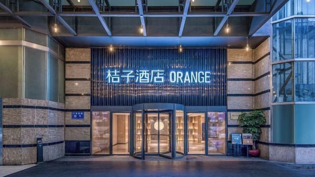 A entrada do hotel Orange com uma porta giratória, uma placa acima da porta e uma planta em vaso à direita.