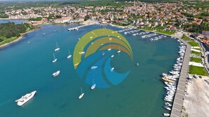 Vista aérea de un pueblo costero con un puerto deportivo, barcos y una extensa zona verde con un logotipo distintivo.