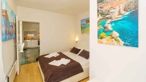 A bedroom with a bed, a painting of a coastal scene, and a view of a kitchen and bathroom. - Holiday apartment with air conditioning (Crikvenica)